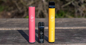 Three vape pens in pink, black, and yellow on a wooden surface.